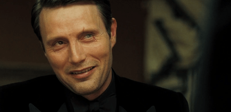 Mads Mikkelsen smiling in a black suit and scar on his eye