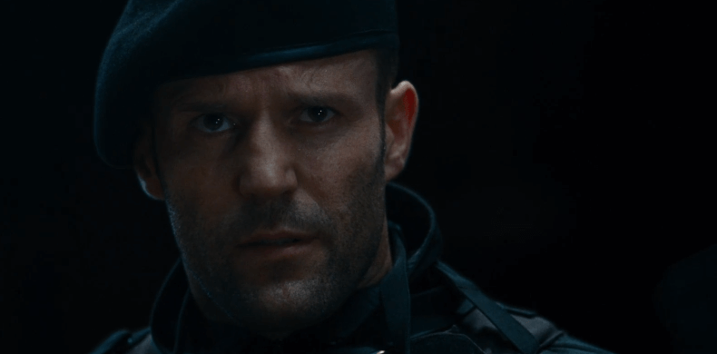 Jason Statham in the dark with a tilted hat looking serious