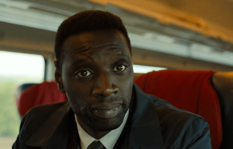Omar Sy looking serious in a suit on a train.