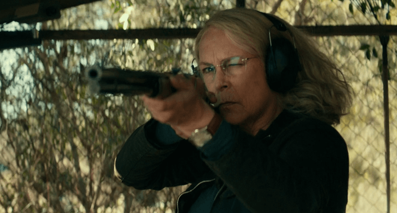 Laurie Strode aiming a weapon at a firing range with glasses and sound dampening headphones.