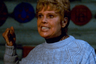 Betsy Miller holding a knife and gritting her teeth while wearing a blue sweater.