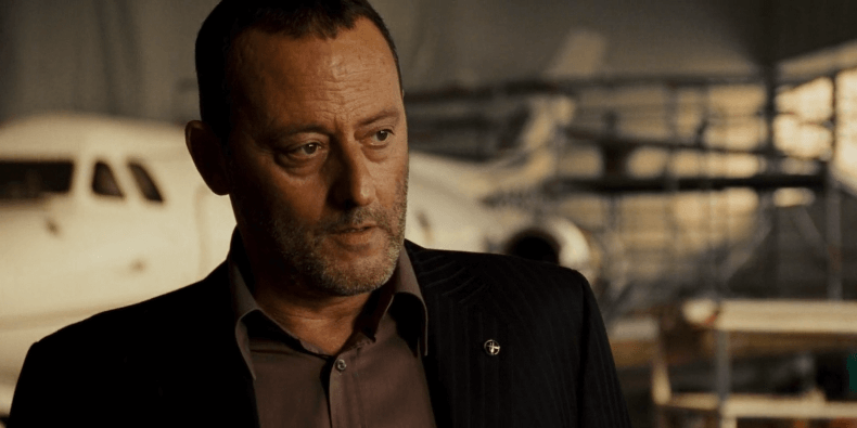 Jean Reno in a black and grey suit in a hangar.