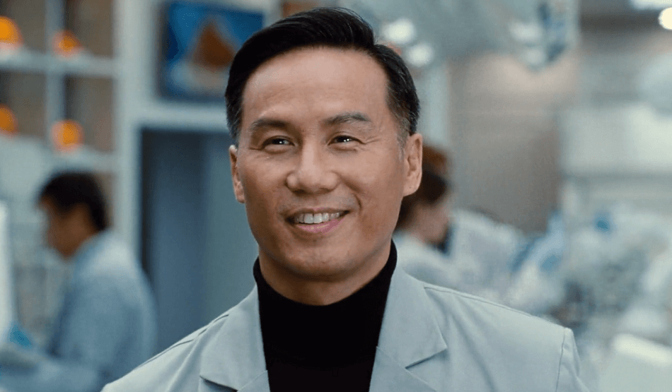 BD Wong smiling brightly inside a lab while wearing a gray suit jacket and black turtleneck sweater.