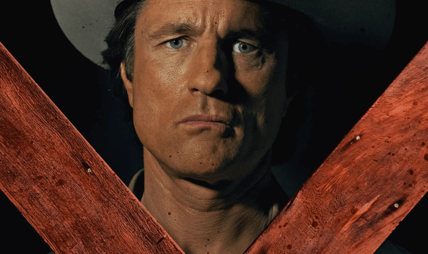 Martin Henderson wearing a cowboy hat and looking dead serious behind wooden planks.