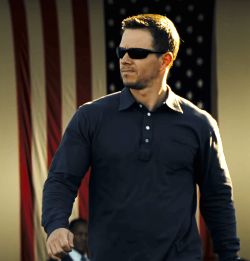 Mark Wahlberg with sunglasses walking with an american flag behind him.
